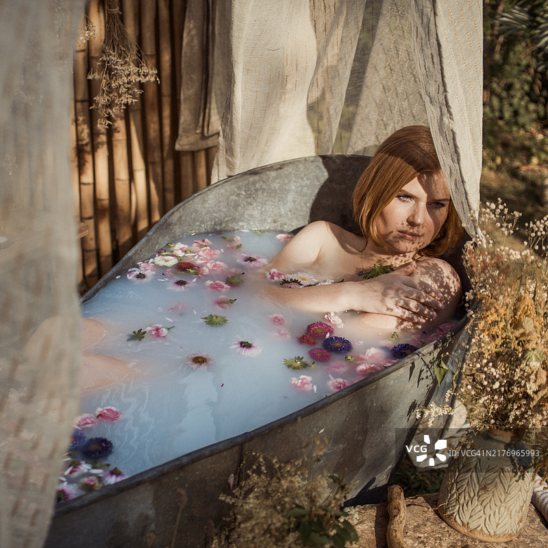 一位红发女性正在花瓣满溢的浴缸中沐浴，浴缸位于花园里。图片素材