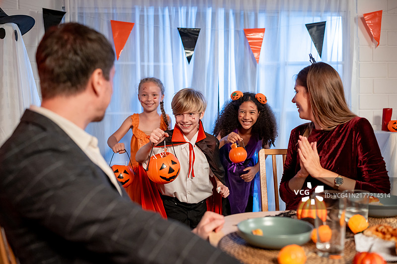 多种族家庭在房子里一起庆祝万圣节派对。一群穿着万圣节服装的孩子拿着不给糖就捣蛋的南瓜篮，在家里节日期间收到糖果图片素材