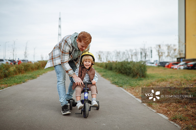 积极育儿：爸爸帮助幼儿儿子学习在秋天骑三轮车图片素材