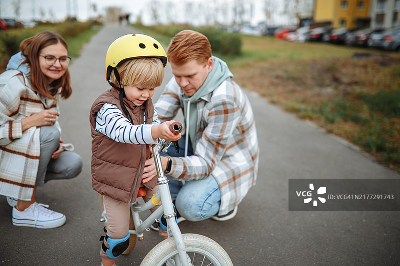 共同育儿——父母共同支持幼儿子的发展，幼儿儿子戴着头盔在住宅区的人行道上一起骑平衡自行车图片素材