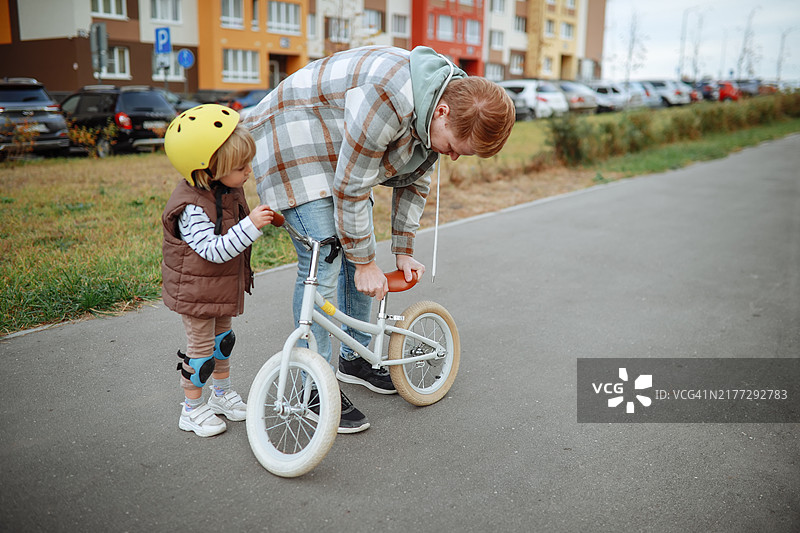 年轻父亲帮助戴黄色头盔的幼儿儿子学习骑平衡自行车图片素材