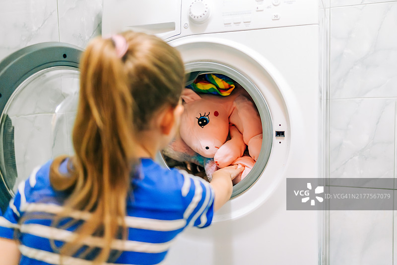 小学女生将一个大的粉色独角兽毛绒玩具放入洗衣机，体现了家务、清洁和责任感图片素材