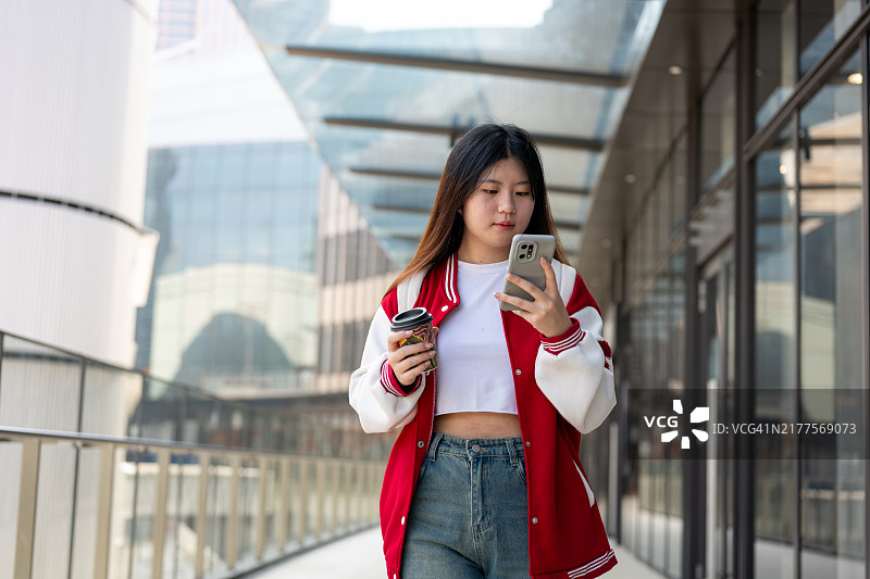一位美丽的年轻Z世代亚洲女性走在人行道上，手里拿着咖啡杯和手机。图片素材