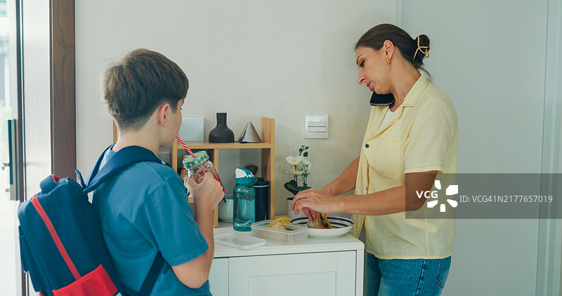 年轻母亲一边准备午餐一边打电话，她的儿子背着背包，在上学前喝着冰沙，重返校园的概念图片素材
