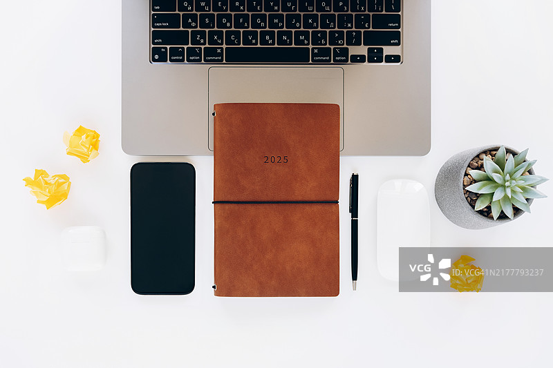 现代工作区，铺有2025年计划本、笔记本电脑、智能手机、无线鼠标、笔、多肉植物和白色桌子上皱巴巴的纸。简约的办公设置，提高工作效率和计划图片素材