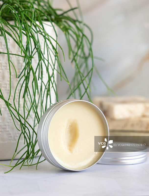 打开的身体乳霜罐放在大理石台面上，旁边有绿色植物特写，化妆品展示模版图片素材
