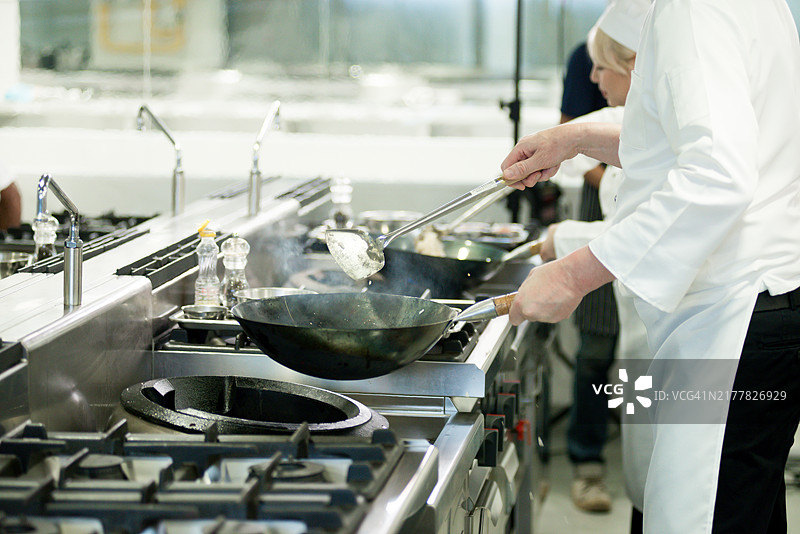 两位多种族厨师在酒店或餐厅厨房忙于烹饪，手持煎锅准备美味的食物，正在烹饪学校工作。这些厨师在高档餐厅厨房工作。图片素材