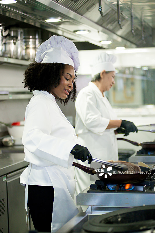 年轻的黑人女学生在现代餐厅的厨房里与她的女老师一起，熟练地烹饪美味多汁的带骨猪排，油锅中冒着热气。学生们在餐厅的烹饪课堂上。图片素材