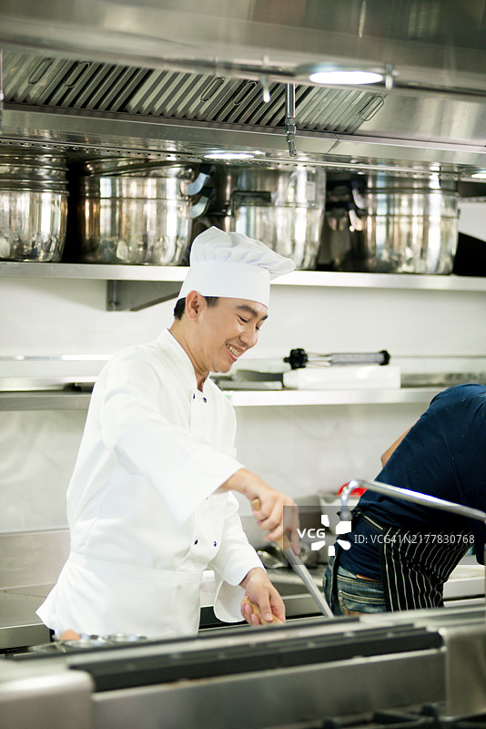 亚洲中年男性厨师在餐厅专业厨房为午餐服务烹饪美食，参加烹饪课程。微笑的亚洲男子手持平底锅，准备在烹饪学校制作美味食物。图片素材
