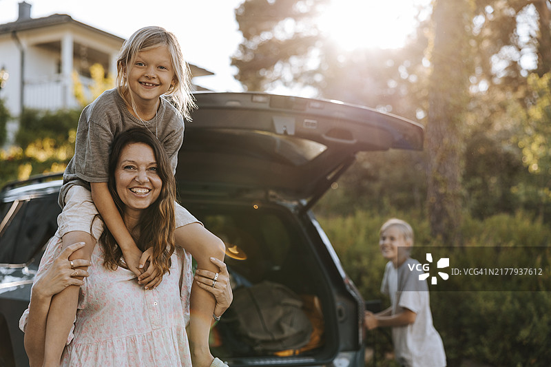 快乐的女人站在汽车后备箱前，儿子坐在肩膀上的肖像图片素材