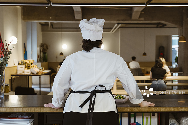 女厨师在厨房岛附近观看坐在餐厅里的顾客的背影图片素材