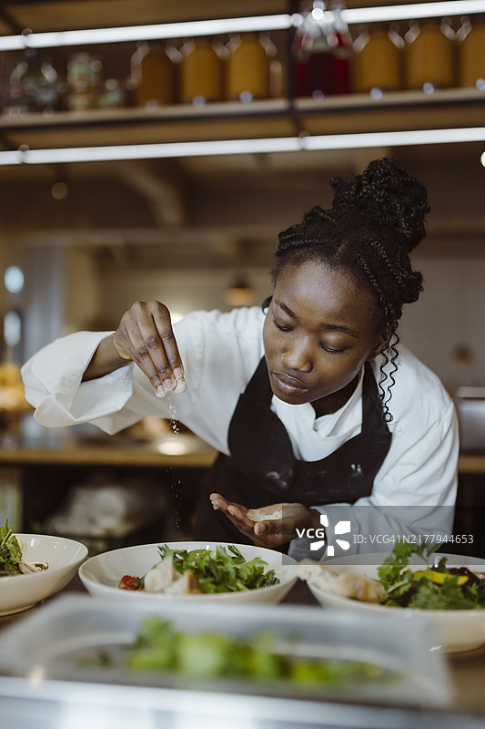 专注的年轻女厨师在餐厅的商业厨房弯腰，在沙拉上撒盐图片素材