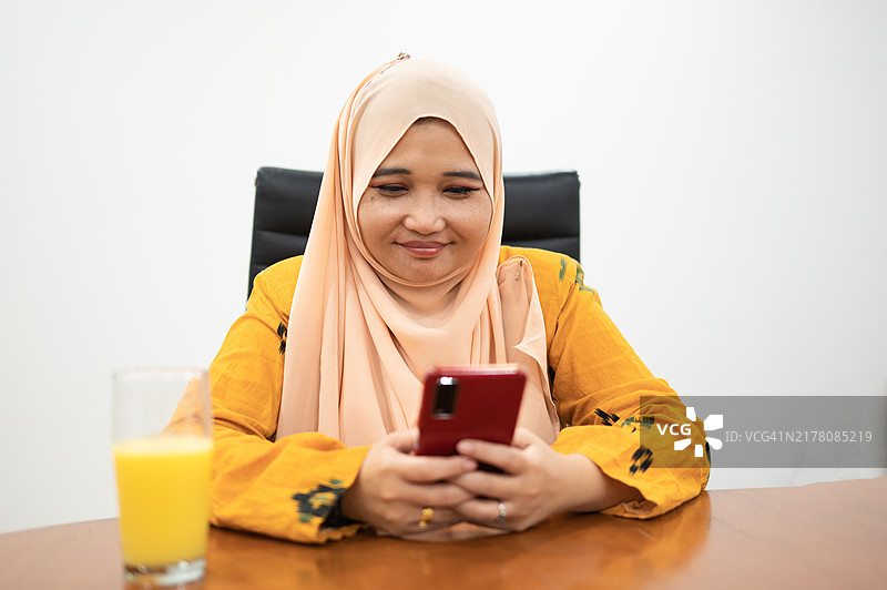 亚洲女性在室内使用智能手机，手边有饮料，面带微笑的照片图片素材