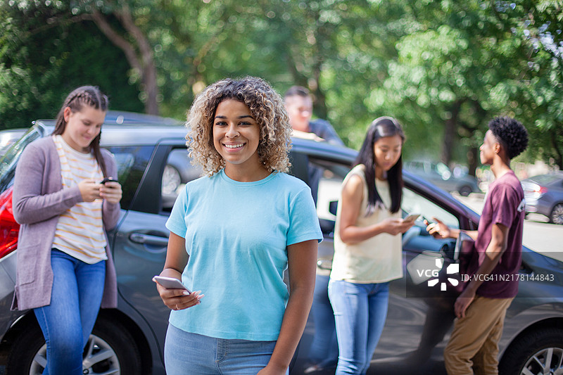 站在停车场汽车前的年轻混血少女与朋友们使用智能手机图片素材