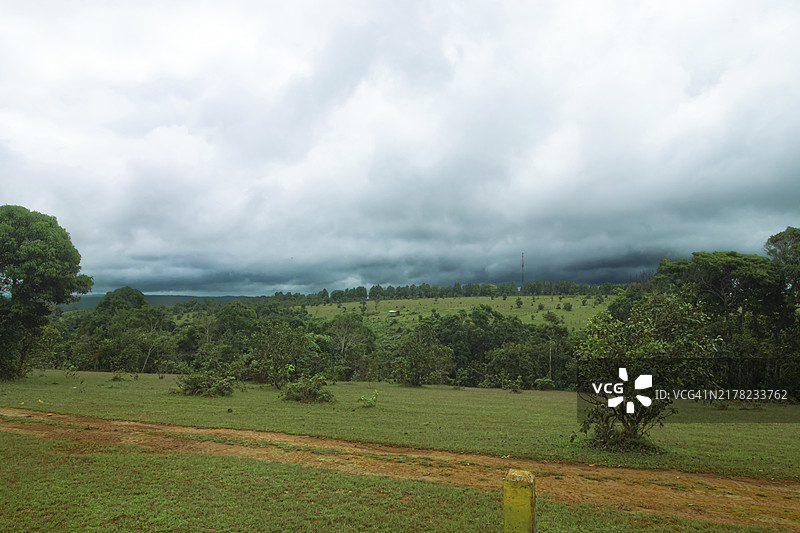 阴天多云天空下的绿色山丘景观，一条土路横穿而过——柬埔寨蒙多基里省，亚洲图片素材