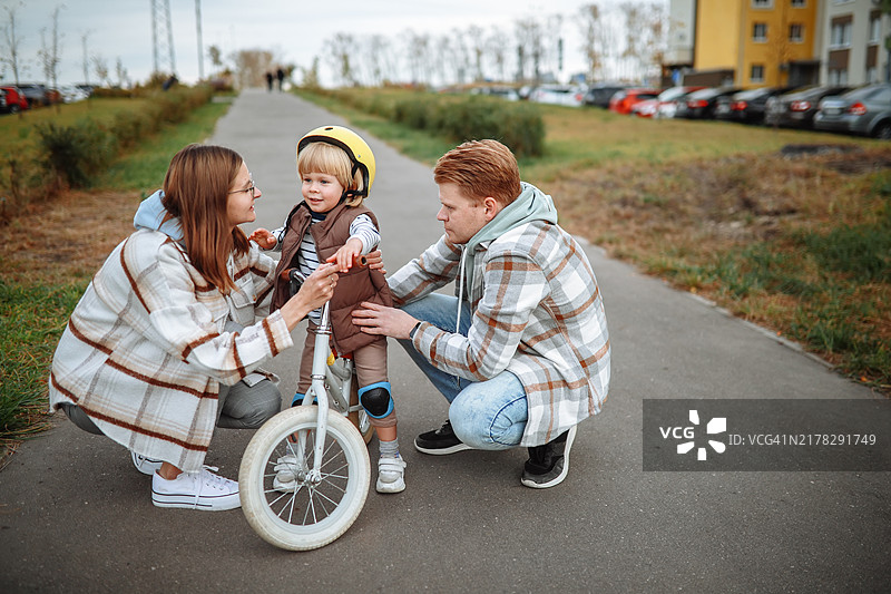 父母合作：妈妈和爸爸一起帮助蹒跚学步的儿子骑平衡自行车图片素材
