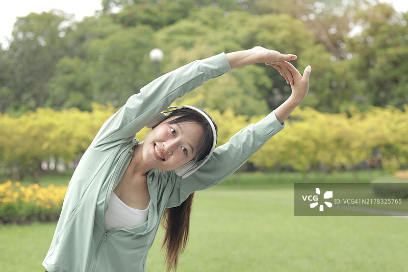 快乐的亚洲年轻女孩在公园户外戴着耳机做轻度运动。微笑的运动女性穿着运动服在夏季绿意盎然的花园中进行身体拉伸。健康的女性在进行个人自我护理的锻炼活动。图片素材