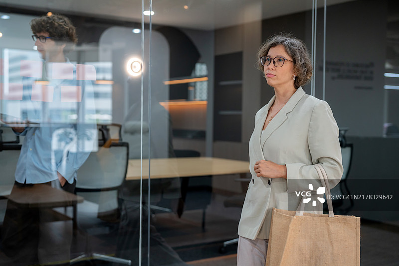 女商人走过现代化的办公室，查看平板电脑上的信息。她透过玻璃墙会议室看去，一群专业人士站在那里，热烈地讨论工作。图片素材