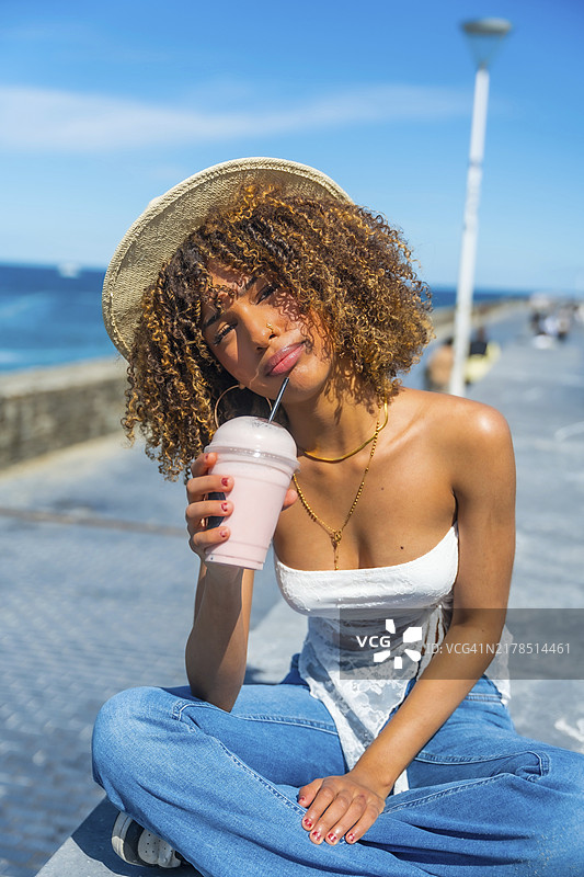 穿着夏装戴着遮阳帽的拉丁裔少女在海边长廊喝奶昔竖版肖像图片素材