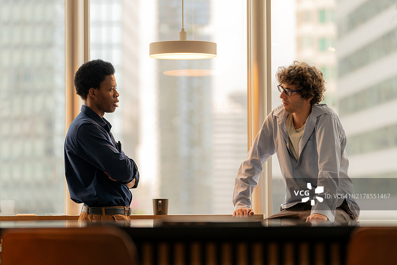 两名商务人士在现代办公室的黄昏中一边喝咖啡一边讨论项目计划。图片素材