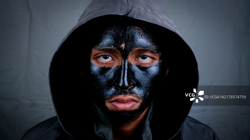正面视图的男子穿着连帽衫，脸上涂有黑色泥浆面膜，背景为空白。图片素材