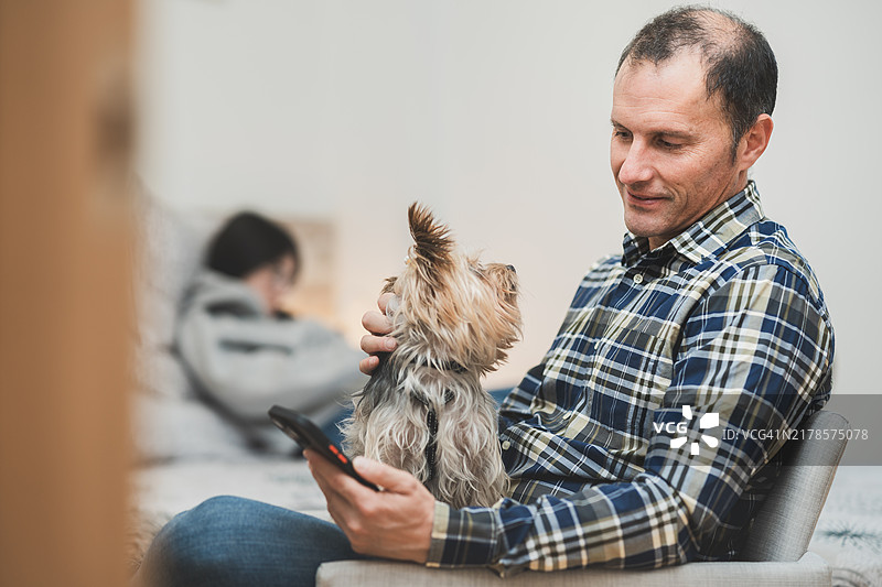 男人和狗在家放松，青少年儿子在背景中使用智能手机图片素材