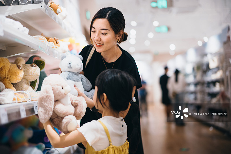 年轻的亚洲母亲和她可爱的女儿在玩具店里快乐地购买儿童软玩具。她们在购物中心一起享受时间和购物。家庭购物和生活方式。零售和消费主义图片素材