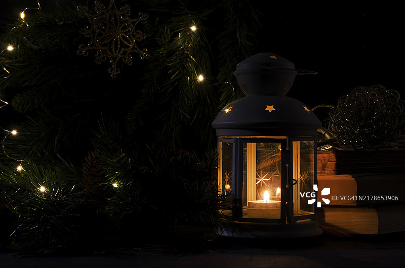 带有燃烧蜡烛的白色灯笼，背景是书籍、圣诞树枝和灯光，夜晚黑暗的画面，圣诞节神秘气氛图片素材