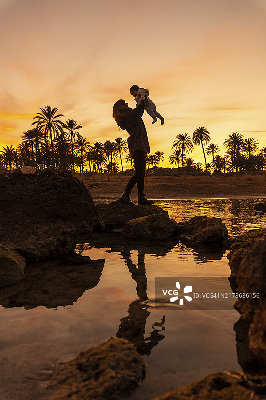 海滩上的家庭生活，年轻母亲和她的宝宝在橙色日落的海边，棕榈树海滩图片素材