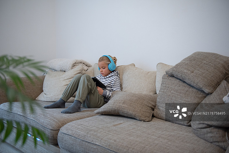 男孩戴着耳机坐在沙发上，在iPad上看电影图片素材