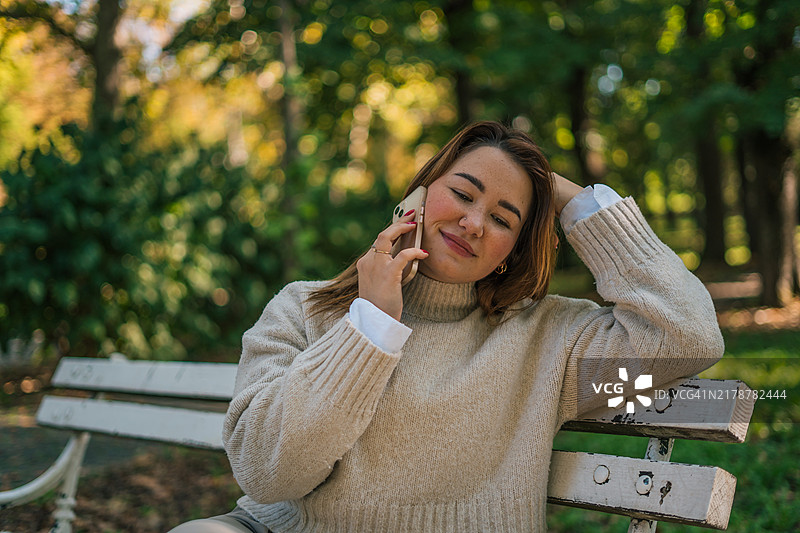 快乐的女士在户外长椅上打电话，拿着塑料杯。都市生活和交流概念图片素材