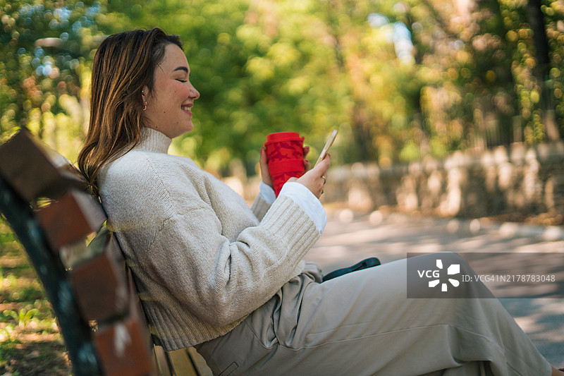 快乐的女士在户外长椅上打电话，拿着塑料杯。都市生活和交流概念图片素材