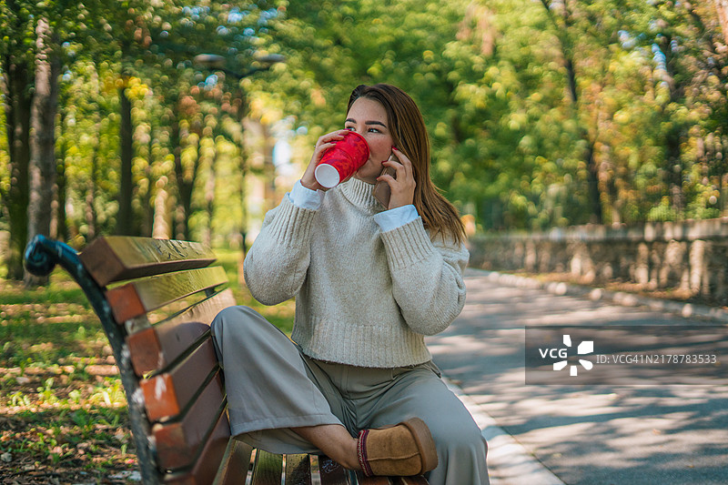 快乐的女士在户外长椅上打电话，拿着塑料杯。都市生活和交流概念图片素材