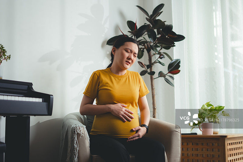 坐在家中扶手椅上感到胃痛的亚洲孕妇图片素材
