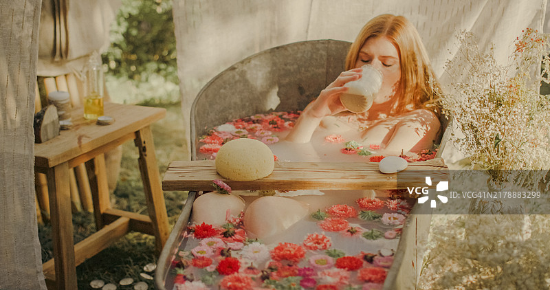 一位红发女性在花瓣满溢的浴缸中喝咖啡，浴缸位于花园里。图片素材