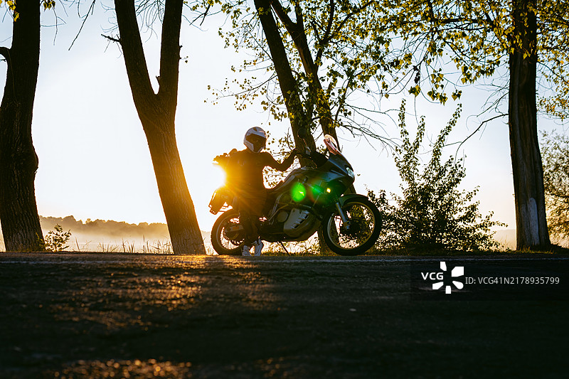 摩托女骑士的剪影，穿着保护服和头盔的女性骑手，骑着大型旅行摩托车，背景是树木和湖泊，夕阳西下的傍晚时分。图片素材
