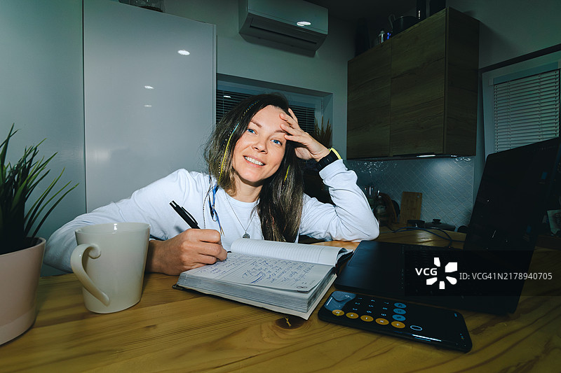 女人在笔记本上写字，旁边有笔记本电脑、咖啡杯和智能手机，营造出一个温馨的夜间家庭办公室环境。图片素材