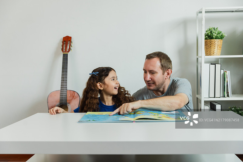 父亲和女儿在家中享受故事时间，一起在厨房桌子上翻阅一本色彩丰富的书。图片素材