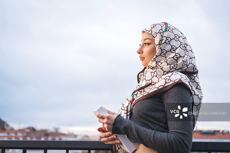 在咖啡店露台上，一位戴着白色面纱的阿拉伯女孩手捧书本，从阳台上眺望城市图片素材