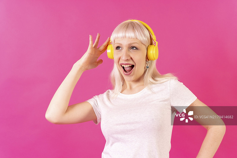 在粉色背景影棚里戴着耳机听音乐微笑的金发白人女孩图片素材