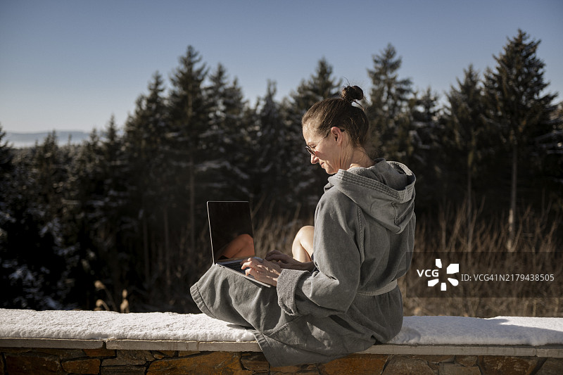 穿着浴袍坐在雪地石墙上用笔记本电脑工作的女人图片素材