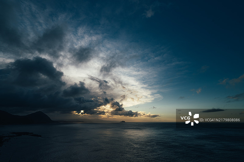 一朵深蓝色的云悬浮在基隆小岛上，带来局部降雨，而远处的天空依然晴朗，映照着日落的色彩。位于台湾新北市。图片素材