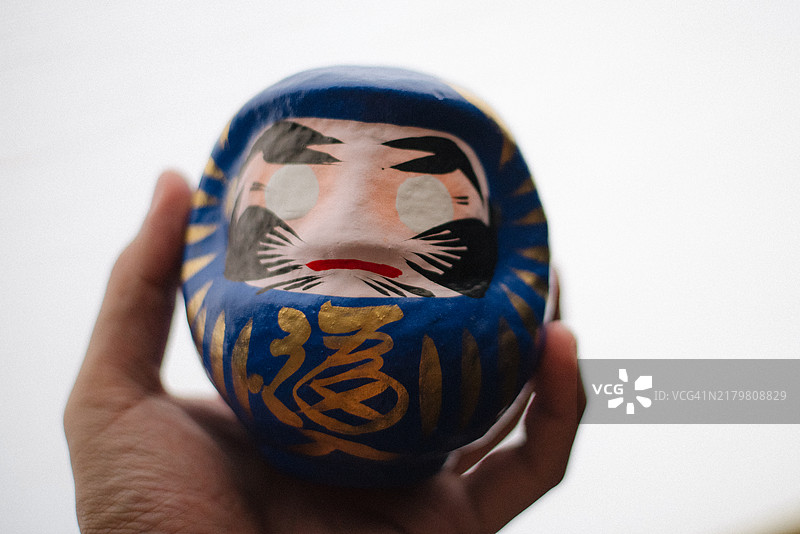 手握蓝色的日本达摩娃娃，这是一种象征毅力和好运的玩偶，以其在跌倒后总能重新站立的韧性而闻名。图片素材