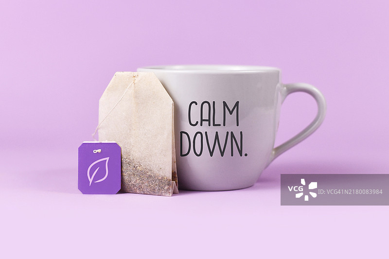 平静草药茶概念：茶包带有标签上的叶子，旁边是茶杯，紫色背景上带有文字“冷静下来”图片素材