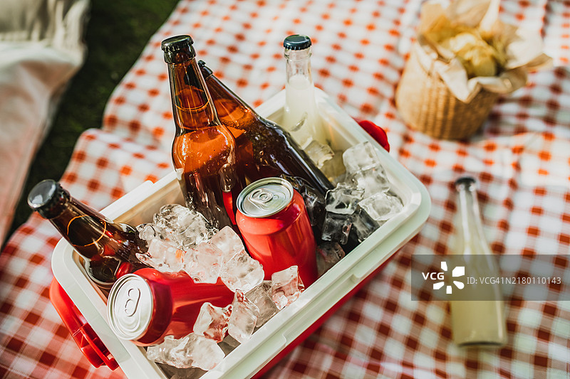 红色塑料冷藏箱，里面放着饮料和冰块，铺在草地上的格子布上图片素材