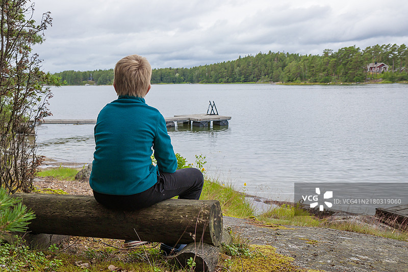 10岁男孩在夏季假期中，坐在海边的木头长椅上休息，期待着背景中的水面和森林，芬兰乡村的宁静，放松，野生自然，海岸。图片素材