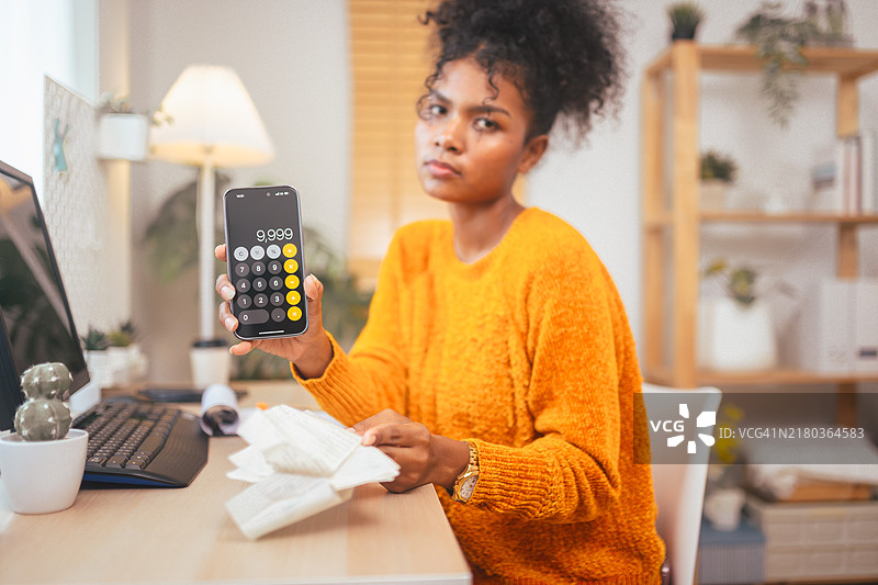 穿着橙色毛衣，拿着计算器和收据，表情担忧，桌上有笔记本电脑，表明存在财务管理或预算问题的女人图片素材