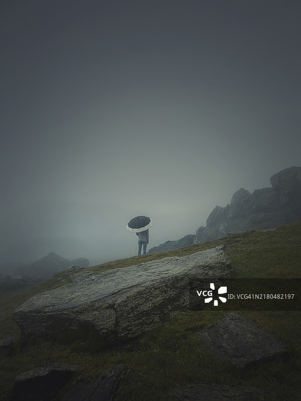 孤独的男人打着伞站在雾气笼罩的岩石山上的背影图片素材