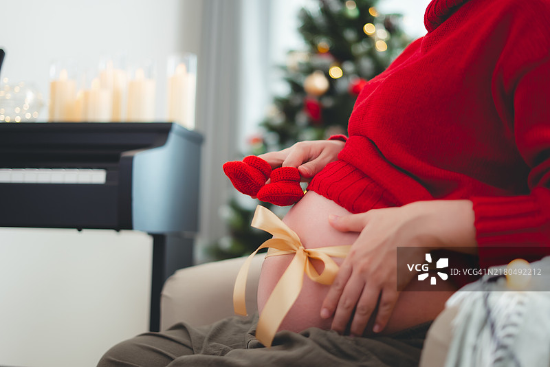 孕妇展示系着蝴蝶结的肚子和婴儿鞋（圣诞树背景）图片素材