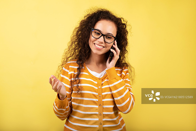 黄色背景下使用手机的深色皮肤女人，视频通讯自拍，录音播客，博主，远程在线工作，智能手机上的对话图片素材